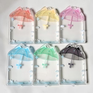 May include: Six rectangular clear acrylic card holders featuring a printed design of a cat-eared umbrella in red, yellow, pink, blue, green, and black. Each holder includes small blue raindrop and star graphics, with a small loop at the top.