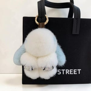May include: A fluffy white and pale blue bunny keyring with rhinestone detailing on the feet. The keyring is attached to a black leather strap and gold ring, with a black bag in the background. The word "STREET" is visible on the bag.