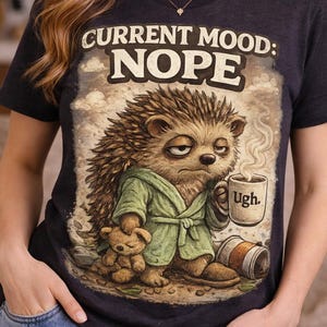 Op de afbeelding: Donkergrijs t-shirt met de tekst "CURRENT MOOD: NOPE" en een cartoonegel in een badjas, met een koffiemok. De egel heeft een chagrijnige uitdrukking en houdt een teddybeer vast.