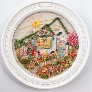 May include: An embroidered artwork in a white frame, showing a cottage scene. The cottage has a green roof, a blue door, and a window covered in flowers. A yellow sun shines above, and a path leads through a garden with colourful flowers.