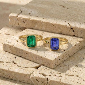 Op de afbeelding: Twee gouden ringen met smaragdgeslepen edelstenen. Eén ring heeft een levendige groene smaragd, de andere een diepblauwe saffier. De ringen zijn gezet in gouden banden en worden getoond op een getextureerd, lichtgekleurd oppervlak.