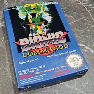May include: A blue Bionic Commando video game box for the Nintendo Entertainment System. The cover features a character in a green and yellow suit holding a weapon, with the Capcom logo, the Nintendo Seal of Quality, and descriptive text.