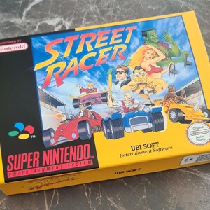 May include: A rectangular video game box for Street Racer on the Super Nintendo Entertainment System. The yellow and black packaging features colourful illustrations of various characters driving race cars, with the Nintendo logo and seal of quality.