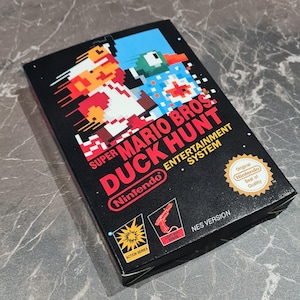 May include: A black Nintendo game box for Super Mario Bros. Duck Hunt. The box features pixelated graphics of Mario and a duck, with the Nintendo logo and "NES VERSION" text. Includes the "Original Nintendo Seal of Quality" sticker.