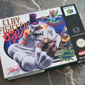 Op de afbeelding: Een Nintendo 64 videogamebox voor "Clay Fighter 63 1/3". De doos toont een cartoon sneeuwpop karakter met een paarse hoge hoed en een bruin, klei-achtig object. De doos toont ook het "Earthworm Jim" karakter.