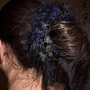 Op de afbeelding: Een persoon met donker haar draagt een scrunchie. De scrunchie is een mix van donkerblauw, groen en zwart, met een getextureerd, pluizig uiterlijk. De scrunchie is gemaakt van een zacht, mogelijk garenachtig materiaal.