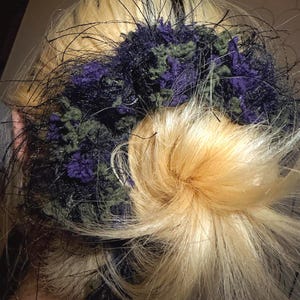 Op de afbeelding: Een handgemaakt haaraccessoire met donkerpaars, groen en zwart garen. Het accessoire is geplaatst tegen blond haar, waarbij het garen een gestructureerd, decoratief element vormt.
