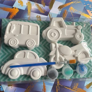 May include: A set of four white plaster vehicle figures including a bus, a tractor, a car, and a motorcycle. The kit includes a blue-handled paintbrush and three small plastic pots containing black, green, and blue paint, all sealed in plastic.