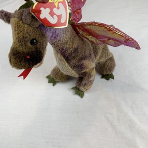 May include: A plush dragon toy with brown and purple fur, black eyes, and a red tongue. It has iridescent red and gold wings and green felt feet. A red and white tag is attached to the dragon's ear.