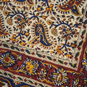 May include: Close-up of a patterned textile with a cream background. The design features intricate floral and paisley motifs in shades of blue, yellow, and red. The border is a deep red with floral patterns.