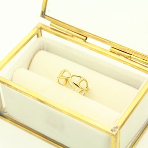 May include: A gold ring with a delicate, openwork design sits inside a glass and brass ring box with a white velvet lining.