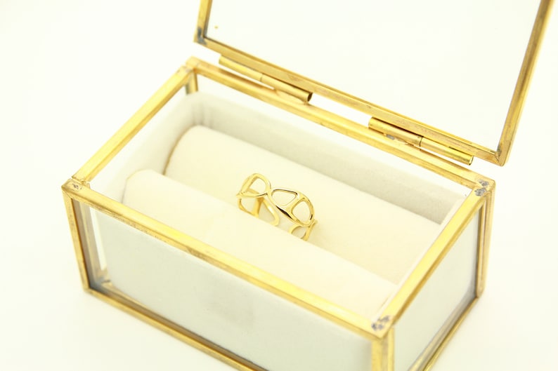 May include: A gold ring with a delicate, openwork design sits inside a glass and brass ring box with a white velvet lining.