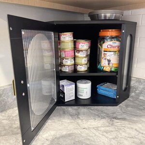 May include: A black corner cabinet with a clear, textured door, open to reveal cat food and treats. Inside are cans of cat food, a jar of Churu treats, and other pet supplies. A stainless steel bowl sits on top.