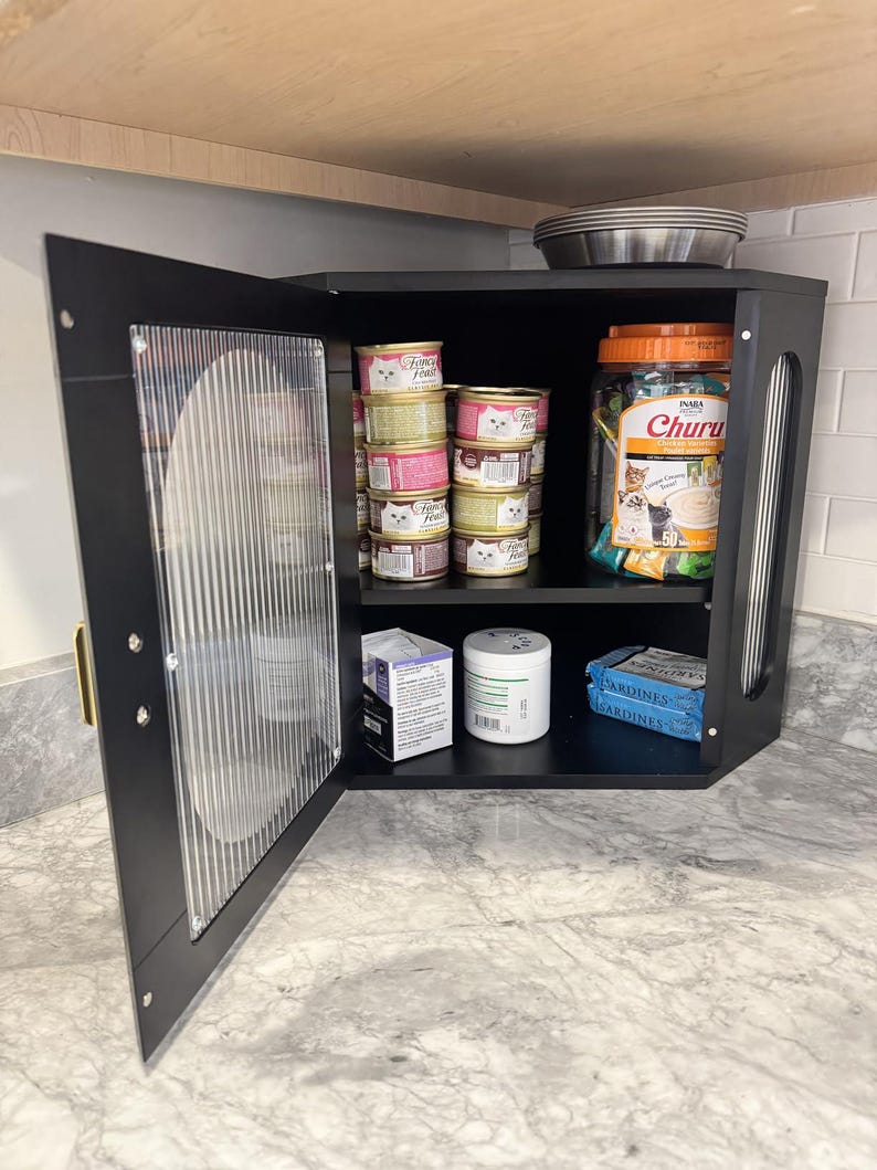 May include: A black corner cabinet with a clear, textured door, open to reveal cat food and treats. Inside are cans of cat food, a jar of Churu treats, and other pet supplies. A stainless steel bowl sits on top.