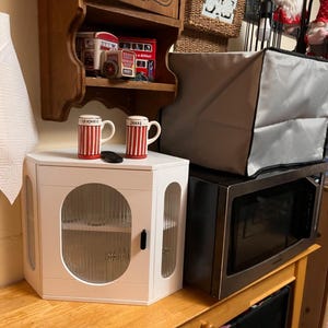 May include: A white corner cabinet with a glass door and a black handle. Two red and white striped mugs sit on top, along with a small black remote. A microwave and a grey cover are behind the cabinet. A roll of paper towels is on the left.