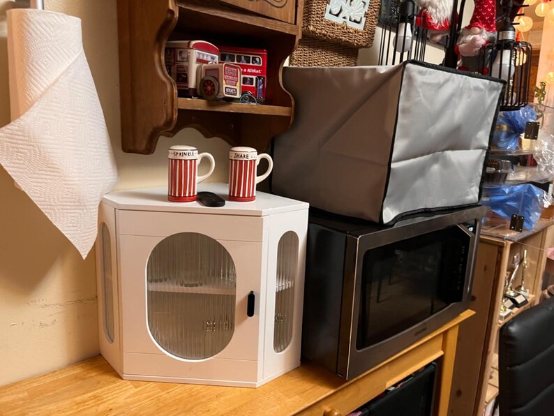 May include: A white corner cabinet with a glass door and a black handle. Two red and white striped mugs sit on top, along with a small black remote. A microwave and a grey cover are behind the cabinet. A roll of paper towels is on the left.