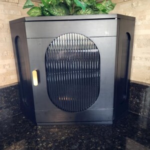 May include: A black cabinet with a rounded front and vertical slats. The cabinet has a gold-coloured handle and is topped with artificial green plants. It is set against a light-coloured brick wall and a dark countertop.