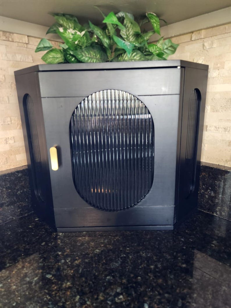 May include: A black cabinet with a rounded front and vertical slats. The cabinet has a gold-coloured handle and is topped with artificial green plants. It is set against a light-coloured brick wall and a dark countertop.