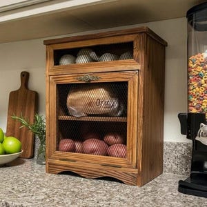 May include: A rectangular wooden storage cabinet with a dark brown finish, featuring a hinged front door with a black wire mesh panel and a decorative metal handle. The interior contains two shelves holding a loaf of bread and several red potatoes.