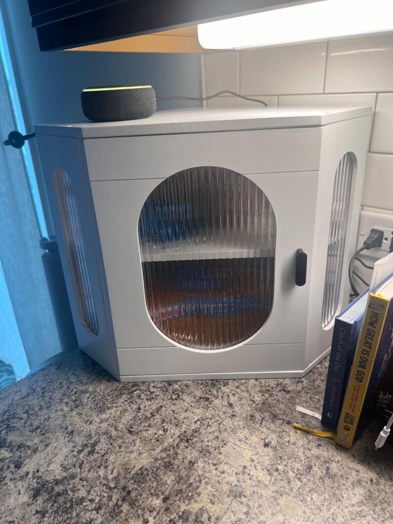 May include: A white, angled cabinet with a clear, textured glass door and side panels. The cabinet has a black handle and contains shelves. An Amazon Echo speaker sits on top. Books are visible to the right.