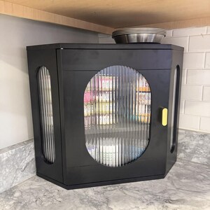 May include: Black hexagonal cabinet with fluted glass door and side panels. The cabinet has a yellow handle and sits on a marble countertop. A stack of metal plates rests on top of the cabinet, beneath a wooden shelf.