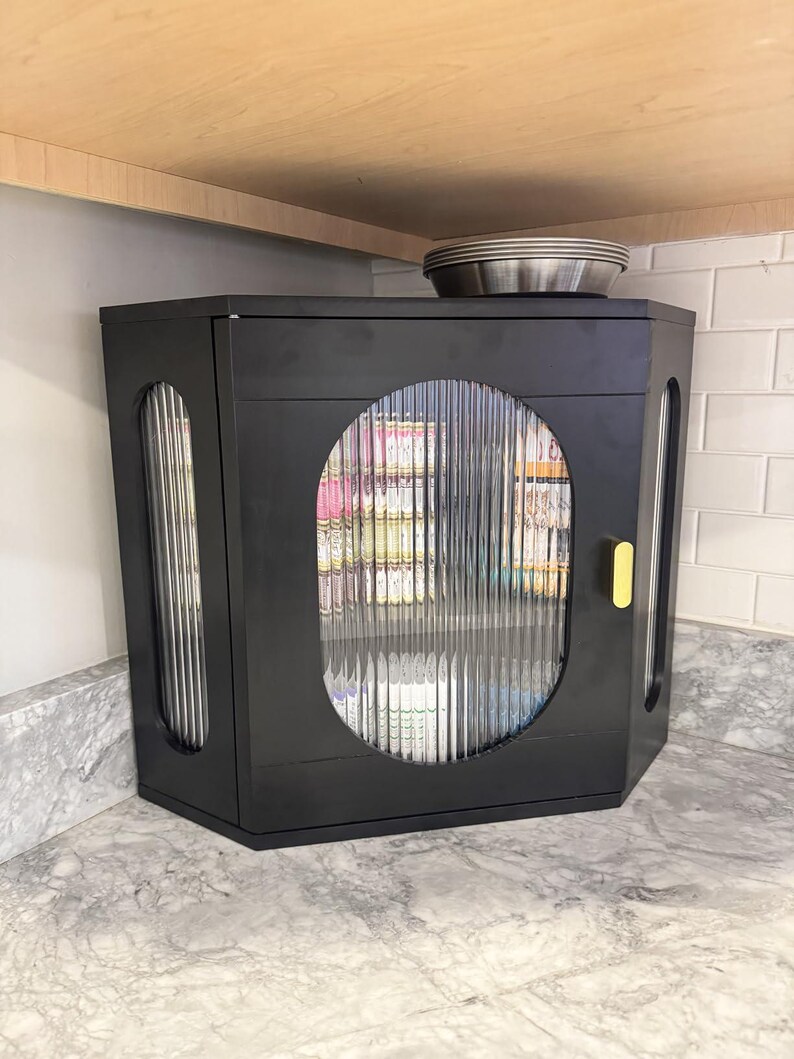 May include: Black hexagonal cabinet with fluted glass door and side panels. The cabinet has a yellow handle and sits on a marble countertop. A stack of metal plates rests on top of the cabinet, beneath a wooden shelf.