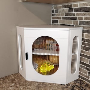 May include: White corner cabinet with a round glass door, ideal for kitchen storage. The cabinet has a black handle and vertical slat details on the sides. The countertop is a speckled brown color, and the backsplash is a mix of brown and gray tiles.