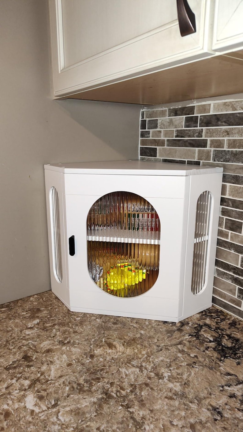 May include: White corner kitchen cabinet with a circular glass door. The cabinet has a black handle and vertical slat detailing on the sides. The countertop is a speckled brown, and the backsplash is a mix of brown and grey tiles.