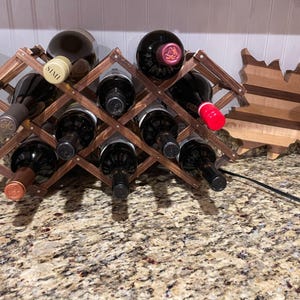 May include: A wooden wine rack holding several wine bottles. The rack is a dark brown color and has a crisscross design. A wooden maple leaf decoration is next to the wine rack. The bottles are various colors, with some labels visible.