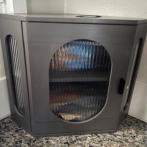 May include: A grey corner cabinet with a textured glass door and side panels. The cabinet has a dark grey finish and a small handle on the side. The cabinet is designed to fit into a corner space.