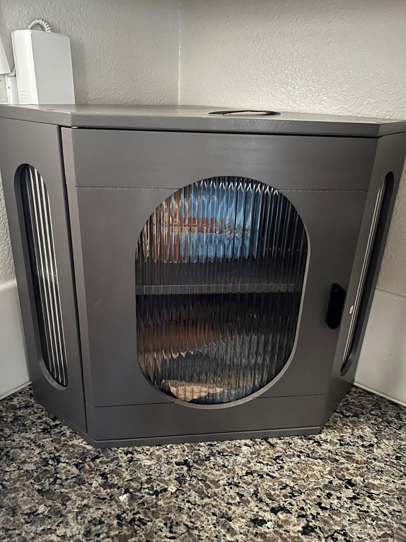 May include: A grey corner cabinet with a textured glass door and side panels. The cabinet has a dark grey finish and a small handle on the side. The cabinet is designed to fit into a corner space.