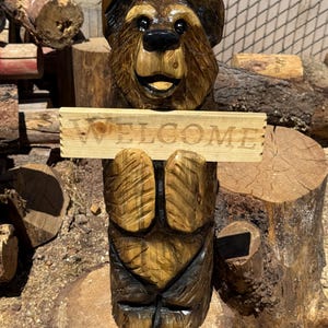 May include: A hand-carved wooden bear sculpture with a dark brown stained finish, featuring a smiling expression and black painted eyes. The bear holds a horizontal light-colored wooden sign with the word WELCOME engraved in capital letters.