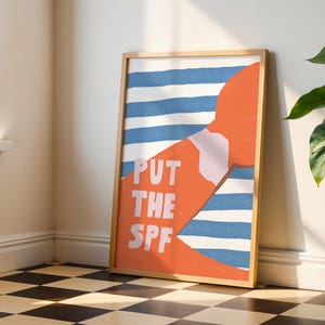 Op de afbeelding: Een ingelijste kunstdruk met een houten frame. Het kunstwerk toont een oranje vorm met een wit en roze gedeelte, tegen een achtergrond van blauwe en witte strepen. De woorden "PUT THE SPF" staan in het wit.
