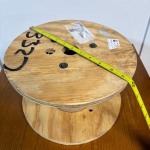 May include: A wooden cable spool with a yellow measuring tape stretched across it. The spool is made of light-colored wood and has a dark number written on the side. The measuring tape shows measurements in inches.