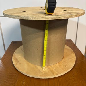 Puede incluir: Una bobina de cable de madera con una cinta métrica amarilla y negra en la parte superior. La bobina está hecha de madera contrachapada y cartón. La cinta métrica muestra una medida de aproximadamente 20 cm.