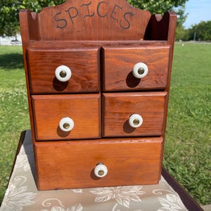 May include: A wooden spice rack with five drawers and white knobs. The word "SPICES" is written across the top. The rack is stained a warm brown colour and sits on a patterned surface.