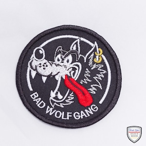 May include: Embroidered patch with a black border featuring a cartoon wolf with a red tongue and a gold earring. The text "BAD WOLF GANG" is in white, arched around the bottom of the design. The patch is on a white background.