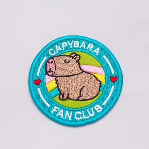 Peut inclure: Écusson brodé avec un capybara de dessin animé à l'intérieur d'un cercle turquoise. Le texte "CAPYBARA FAN CLUB" est arqué autour du capybara. Le capybara est marron avec un nez rose et se détache sur un fond vert et arc-en-ciel. Deux cœurs rouges de chaque côté.