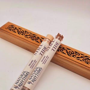 May include: Two tubes of incense sticks with the text "STILL TIDE" and "NATURAL RHYTHMS" are displayed next to a wooden incense holder. The tubes are labeled "ORGANIC SANDALWOOD, AMBER, AGARWOOD, CLOVE". The wooden holder has a decorative carved design.