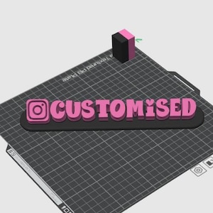 Pode incluir: Uma placa impressa em 3D com o logotipo do Instagram e a palavra "CUSTOMISED" em rosa, sobre uma base preta. Um pequeno bloco preto e rosa está posicionado acima da placa em uma superfície texturizada.