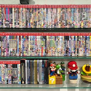 May include: A display of Nintendo Switch game cases with red spines, arranged on glass shelves. The cases feature colourful game art. Mario character figurines are on the bottom shelf.