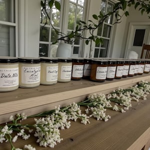 Może przedstawiać: Prezentacja świec w szklanych słoikach z czarnymi wieczkami i białymi etykietami. Etykiety zawierają napisy "Date Nite", "Eucalyptus Mint", "Basil Sage", "White Lilac" i "Lemon Citrus". Świece ustawione są na drewnianej powierzchni.