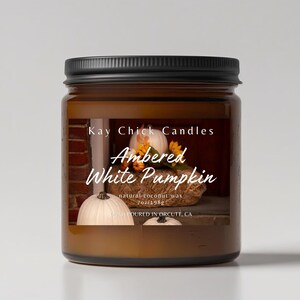 May include: A brown glass candle jar with a black lid and a label featuring the text Kay Chick Candles, Ambered White Pumpkin, and natural coconut wax 7oz/198g. The label displays an image of white pumpkins and autumn flowers.