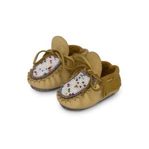 May include: A pair of tan leather baby moccasins. The moccasins have a beaded design on the vamp with colorful beads. They feature a fringe detail on the heel and leather laces.