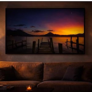 May include: A framed landscape print depicting a sunset over a lake with a wooden pier. The sky transitions from deep blue to vibrant orange and yellow. Dark mountains are silhouetted against the colorful sky. The print is displayed above a brown sofa.
