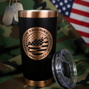 May include: Black and copper-colored insulated tumbler with the text "Operation Enduring Freedom 2001-2021" in a circular design. The tumbler has a clear lid and is set against a camouflage background with an American flag.