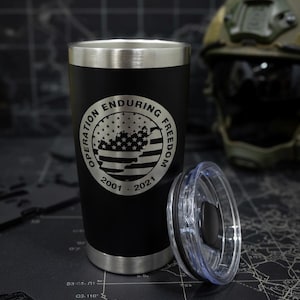 May include: Black tumbler with a silver rim and base, and a clear lid. The tumbler features a circular emblem with the text "OPERATION ENDURING FREEDOM" and a design of the American flag. The years "2001 - 2021" are also displayed.