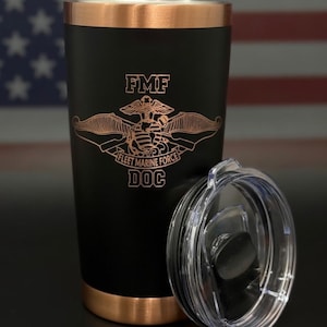 Op de afbeelding: Zwarte beker met een koperkleurige rand en bodem, en een doorzichtig deksel. De beker heeft de tekst "FMF" boven een embleem en de woorden "FLEET MARINE FORCE DOC". De achtergrond bevat een Amerikaanse vlag.