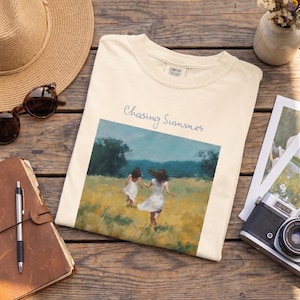 Puede incluir: Camiseta color crema con las palabras "Chasing Summer" y una pintura de dos personas corriendo por un campo. La imagen también muestra un sombrero de paja, gafas de sol, un diario, un bolígrafo, una cámara y un jarrón de flores sobre una superficie de madera.