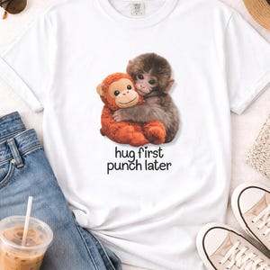 Może przedstawiać: Biała koszulka z grafiką przedstawiającą dwie obejmujące się małpy, jedną pomarańczową i jedną brązową. Pod spodem wydrukowano napis "hug first punch later". Koszulka jest zestawiona z niebieskimi dżinsami i trampkami.